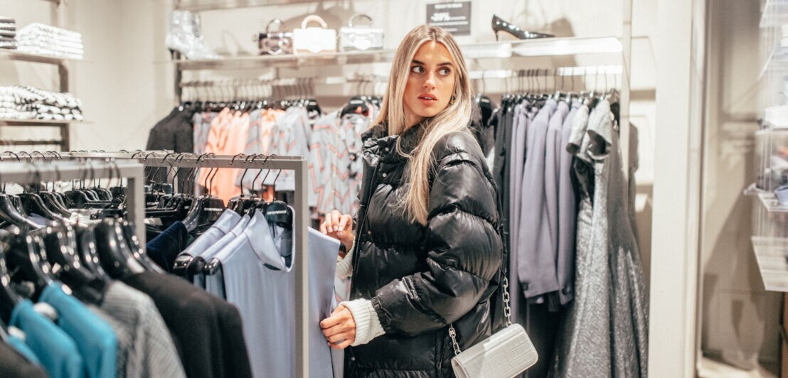 Eine Frau in schwarzer Pufferjacke zwischen Shoppingständern, auf denen Damenbekleidung hängt