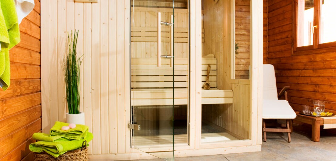 Holzsauna mit Glastür und Fenster, daneben stehen Handtücher sowie ein Sessel und eine Erfrischung bereit