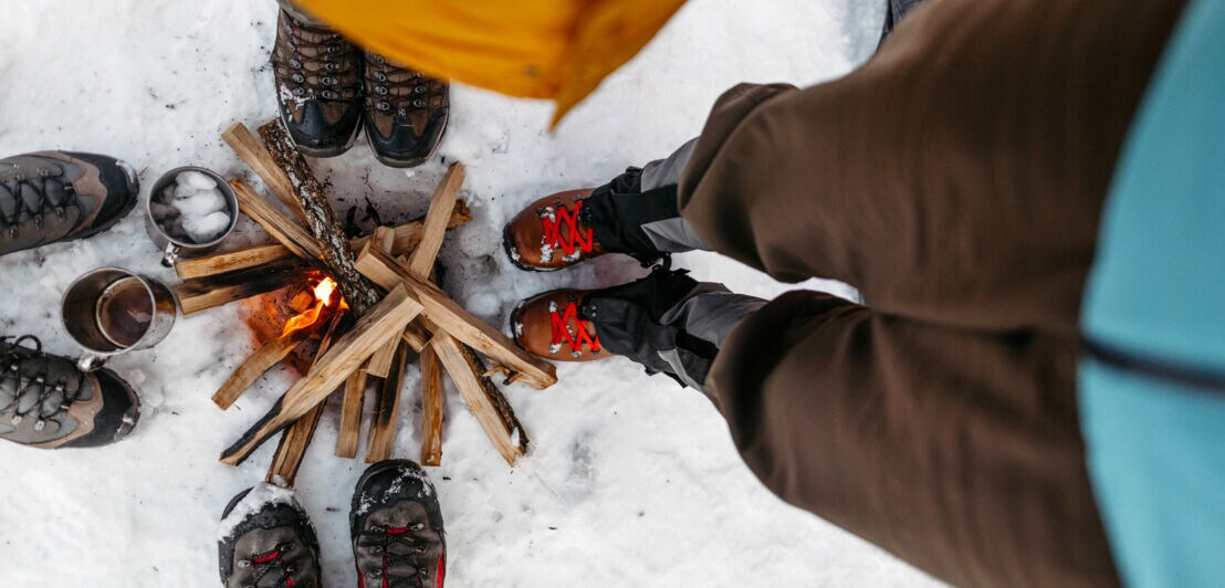 Die Schuhe von vier Personen, die im Schnee um ein Feuer stehen, von oben fotografiert