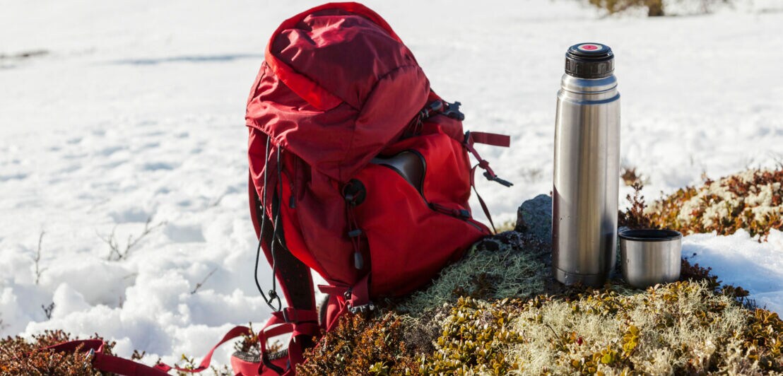 Ein Rucksack und eine Thermoskanne, im Hintergrund Schnee