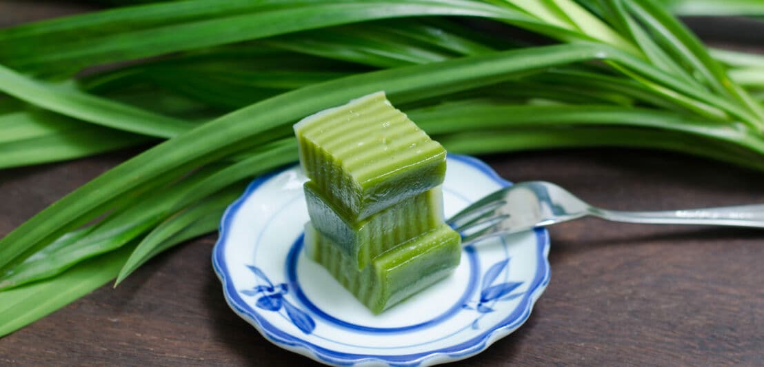 Ein kleiner Teller mit einem grünen Dessert, daneben frische Pandanblätter