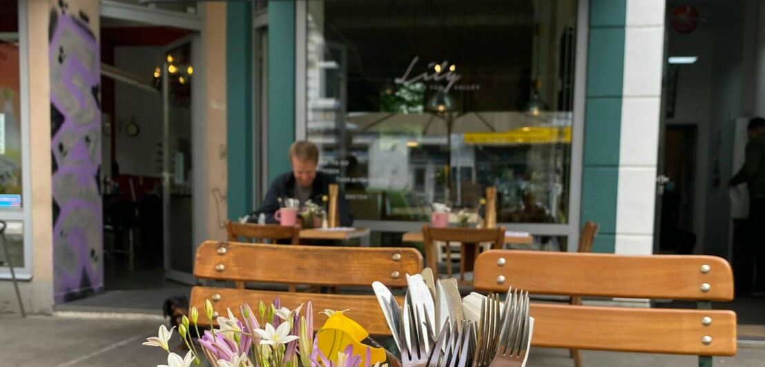 Ein Mann sitzt an einem Tisch vor einem Café im bestuhlten Außenbereich, im Vordergrund Blumen und Besteck auf einem Tisch