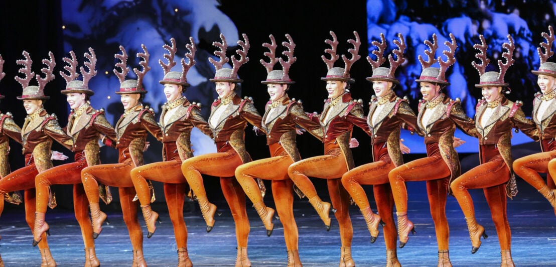 Aufgereihte Tänzerinnen in Elch-Kostümen tanzen auf einer Bühne mit winterlicher Kulisse