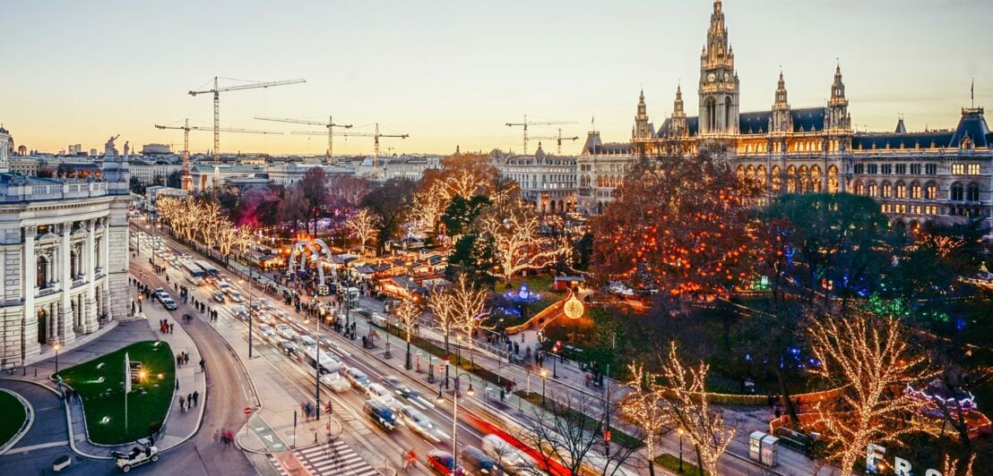 Panoramablick auf das beleuchtete Wiener Rathaus mit Weihnachtsmarkt auf dem großen Vorplatz, davor eine mehrspurige Straße