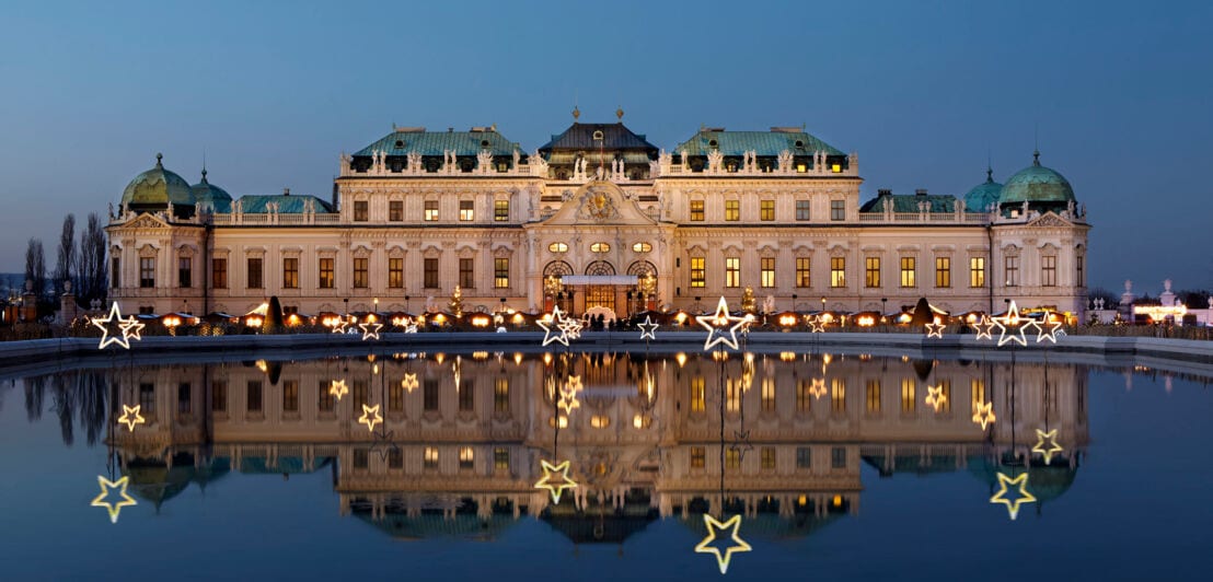 Das erleuchtete Wiener Barockschloss Belvedere mit Weihnachtsmarkt spiegelt sich in der Wasseroberfläche in der Abenddämmerung