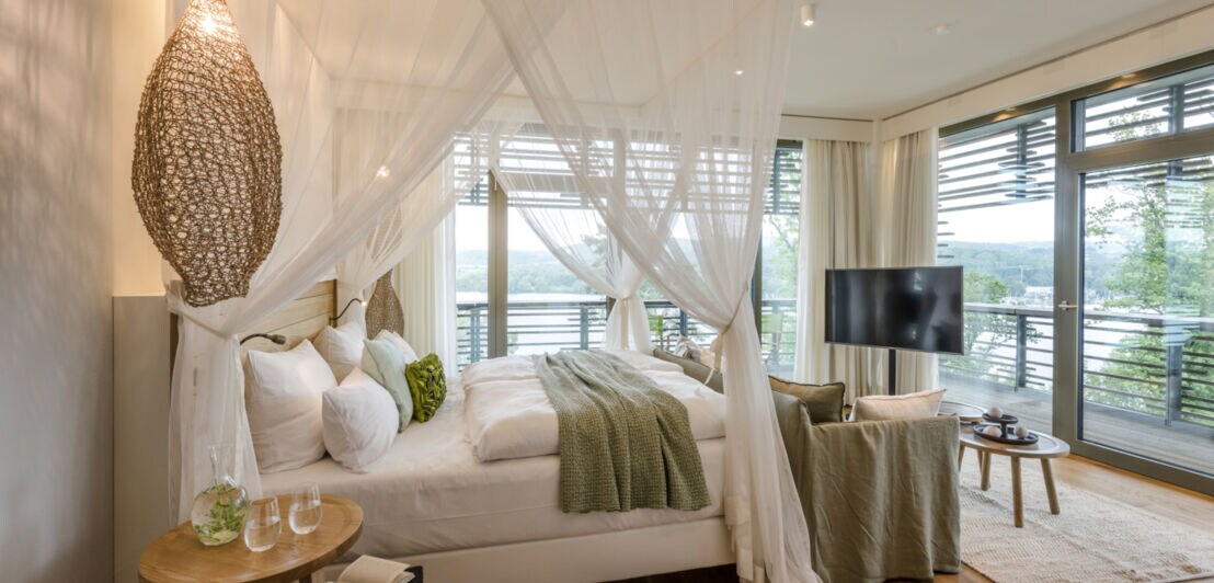 Innenaufnahme eines hellen, modernen Hotelzimmers mit Doppelbett und Panoramafenstern mit Blick in die Natur