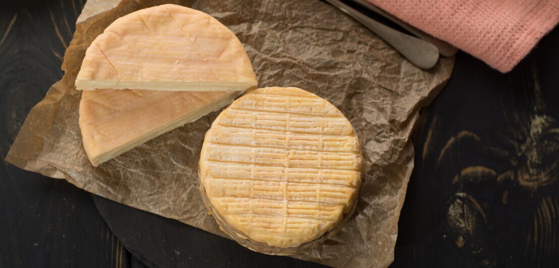 Käsesorte Munster, ein ganzer Käse sowie ein aufgeschnittener liegen auf einem Papier, daneben eine Stoffserviette und Besteck