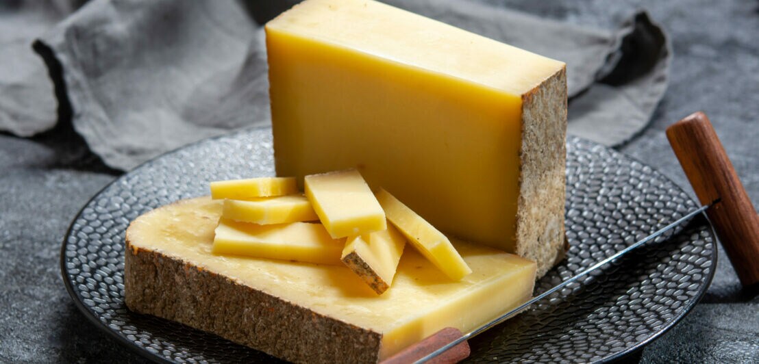 Ein großes Stück Comté-Käse auf einem dunklen Teller, teilweise in Stücke geschnitten, daneben eine Stoffserviette und ein Käsemesser