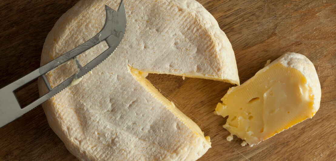Runder Käse auf einer Holzunterlage, ein Stück ist herausgeschnitten und liegt daneben samt Käsemesser