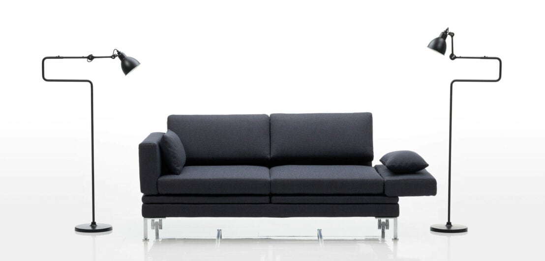 Ein geradliniges, ausklappbares Sofa mit anthrazitfarbenen Bezug, daneben zwei schwarze Stehleuchten im industriellen Design vor weißem Hintergrund