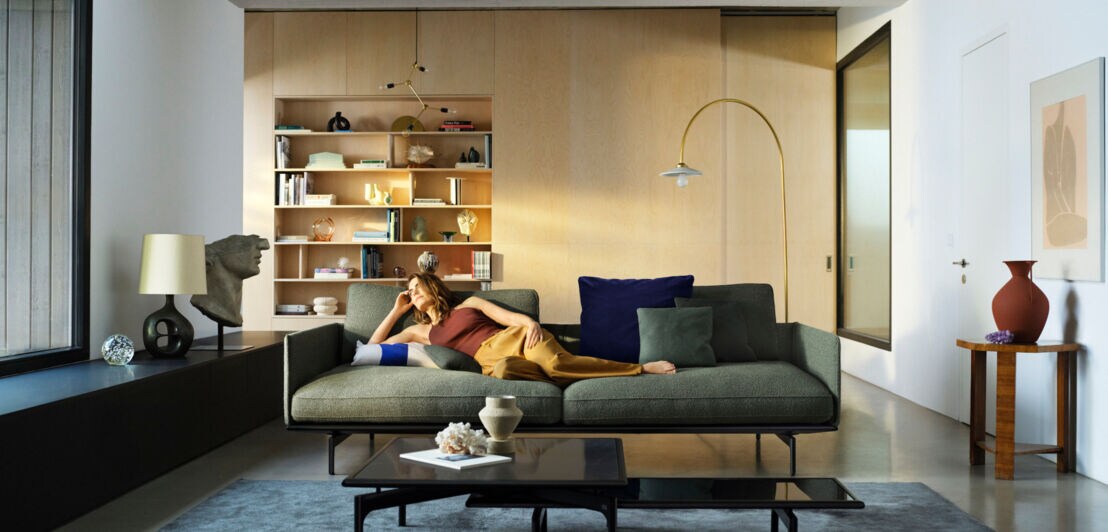 Modern eingerichtetes Wohnzimmer mit Designobjekten, im Zentrum steht ein grünes, geradliniges Sofa, auf dem eine junge Frau liegt