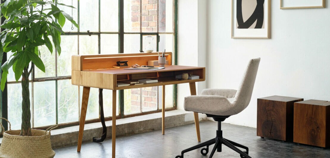 Ein Sekretär aus Holz und ein gepolsterter Schreibtischstuhl auf Rollen stehen in einem modern eingerichteten, hellen Raum mit bodentiefen Fenstern mit Loft-Charakter