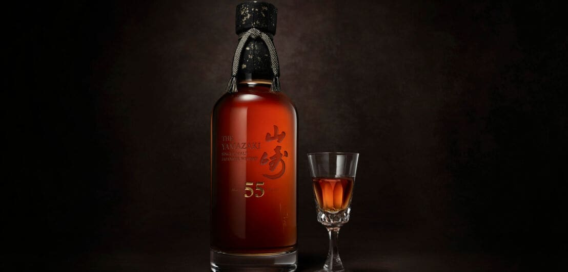 Eine Flasche des Whiskys Yamazaki 55 mit schwarzem Verschluss neben einem hochstieligen Whiskey-Glas vor schwarzem Hintergrund