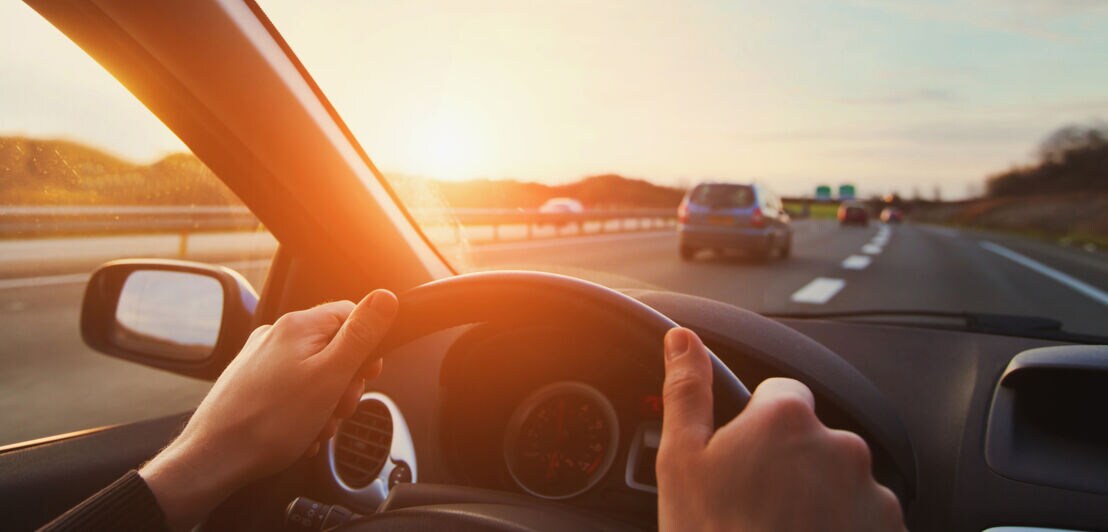 Zwei Hände umfassen ein Autolenkrad, der Blick durch die Frontscheibe zeigt neben der Autobahn auch eine karge Landschaft bei Sonnenuntergang
