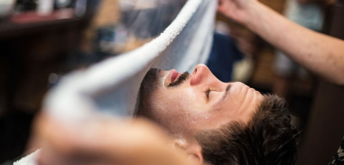 Ein Mann mit geschlossenen Augen in einem Barberstuhl, sein Gesicht wird gerade mit einem warmen, feuchten Handtuch bedeckt