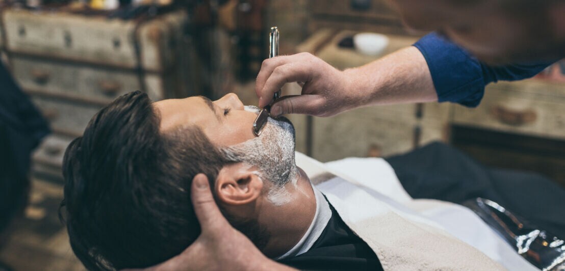 Ein Barber rasiert seinen Kunden mit einem klassischen Rasiermesser den Bart