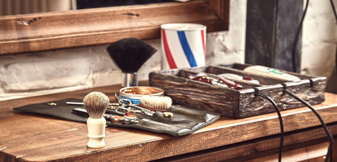 Auf einer Holzkommode unter einem Spiegel liegen klassische Barbertools wie Rasiermesser- und Pinsel