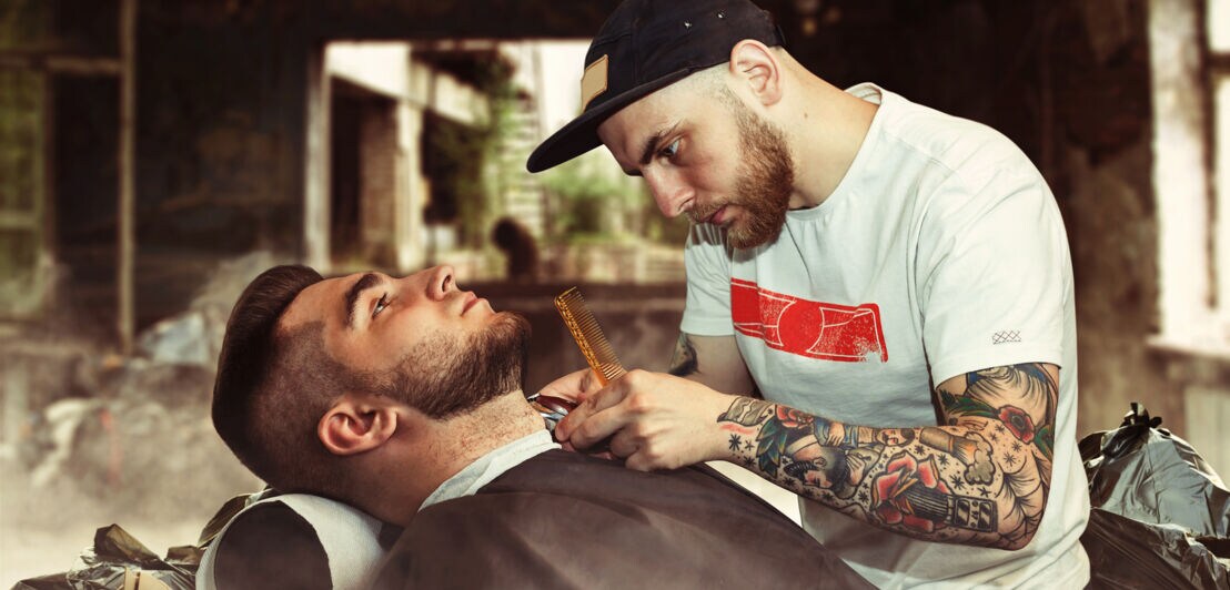 Ein Mann liegt auf einem Friseurstuhl und lässt sich von einem tätowierten Barbier in einem alten Fabrikgebäude den Bart trimmen