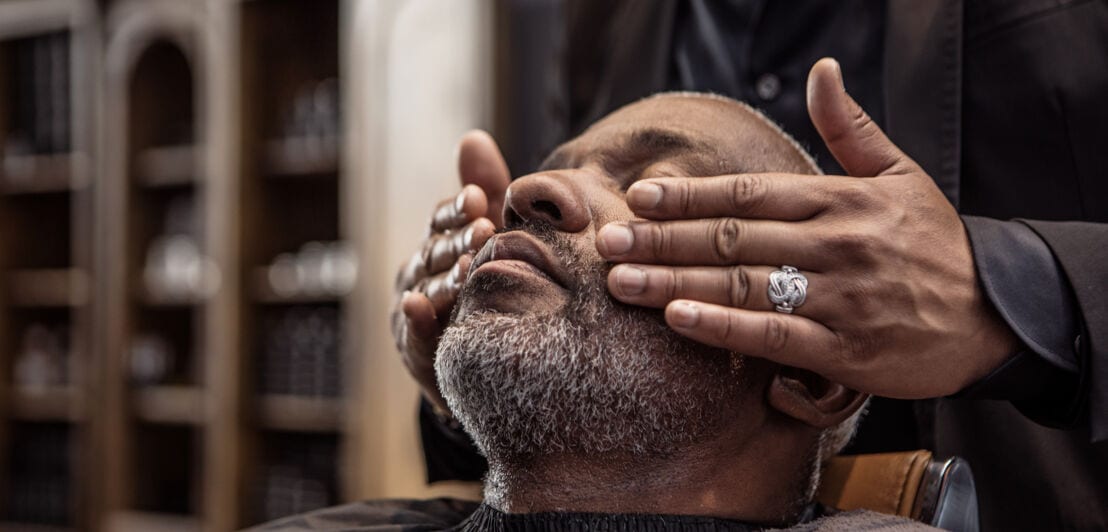 Ein Mann mit frisch getrimmten Bart sitzt mit geschlossenen Augen auf einem Barbersessel und bekommt eine Gesichtsmassage