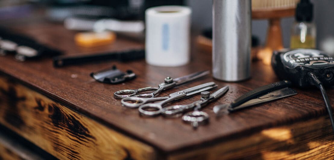 Barber-Zubehör wie drei verschiedene Scheren, Rasiermesser, elektrischer Trimmer und eine silberne Flasche auf einem Holztisch