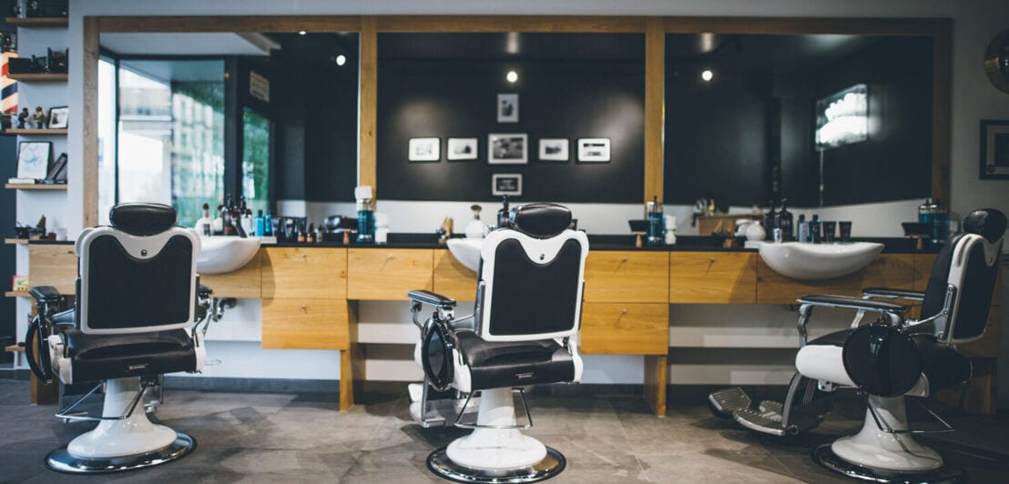 Drei moderne Friseursessel vor Waschbecken und Spiegel in einem Barbershop