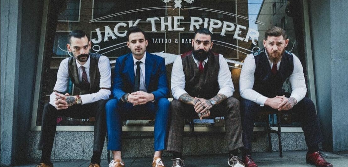 Vier Barbiere in klassischer Herrenkleidung sitzen vor einem Barbershop