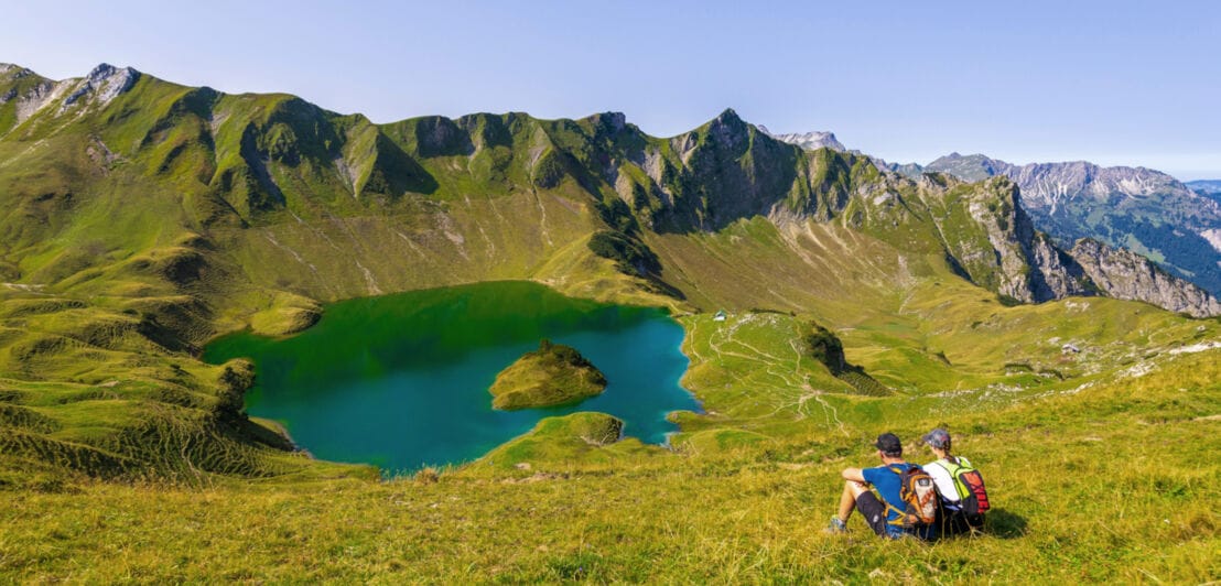 Zwei Wandernde sitzen mit Rucksäcken auf der Wiese vor einem Bergsee