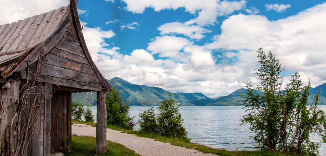 Ein Holzhaus an einem Weg am Ufer eines Sees mit Bergen im Hintergrund