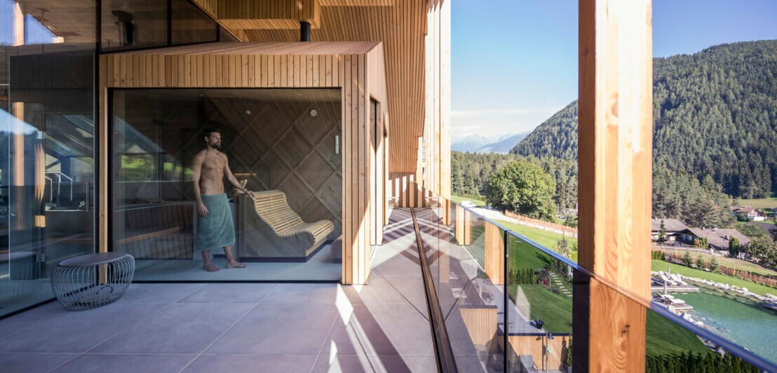 Ein Mann im Badehandtuch steht in einer modernen Holzsauna mit Glasfront auf einer großen Terrasse mit Blick auf eine Berglandschaft