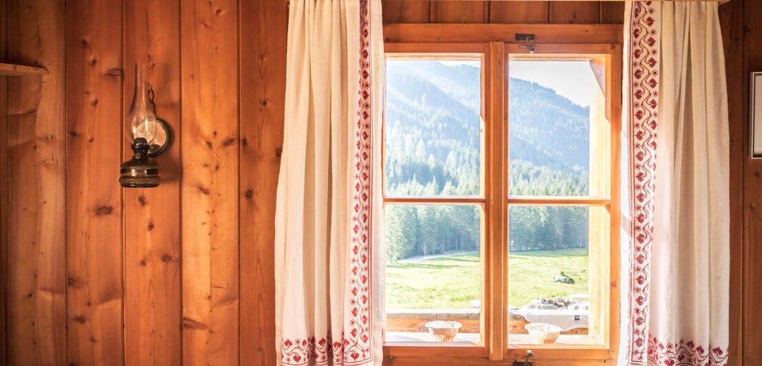 Blick aus einer Holzhütte durch ein Fenster mit Gardinen in die grüne Berglandschaft