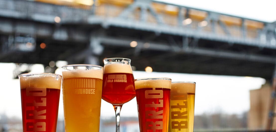 Fünf Biergläser mit unterschiedlichen Inhalten stehen auf einem Tresen, im Hintergrund eine Bahnlinie