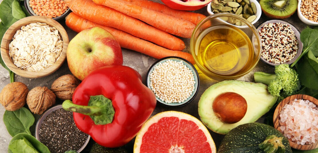 Aufsicht unterschiedlicher Obst- und Gemüsesorten neben kleinen Schalen mit Körnern, Hülsenfrüchten und Öl