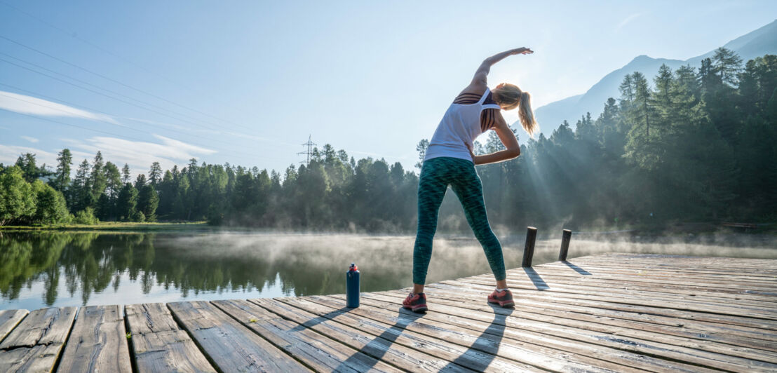 Eine junge Frau in Sportkleidung dehnt ihren Körper auf einem Holzsteg an einem See