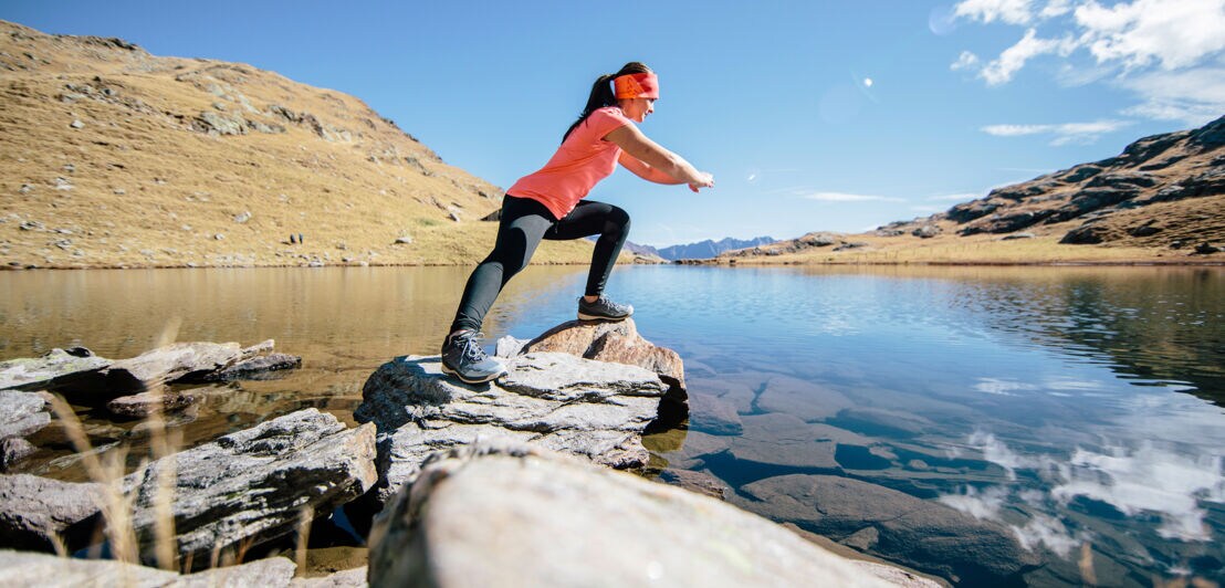 Eine Frau in Sportkleidung steht auf Steinen im Wasser und dehnt sich