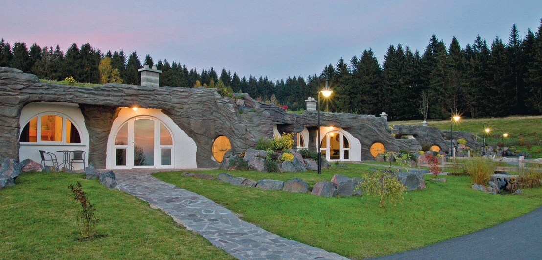 Eine Ferienanlage mit organisch-geformten Behausungen mit Schieferdeckung und begrünten Vorgärten, im Hintergrund Waldlandschaft