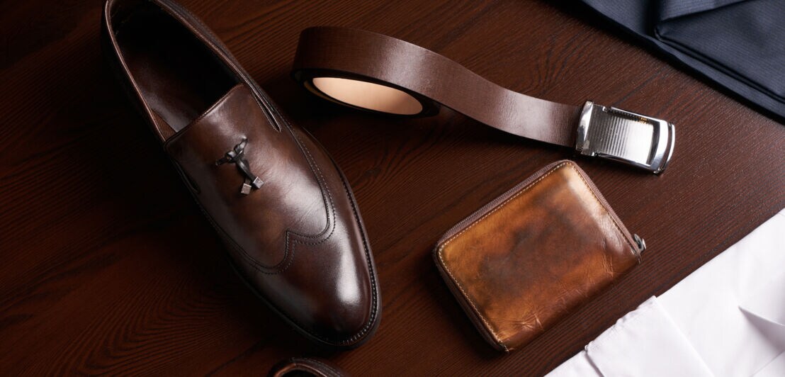 Ein brauner, eleganter Lederschuh für Männer mit einem passendem Gürtel neben einem Portemonnaie auf einem Holztisch