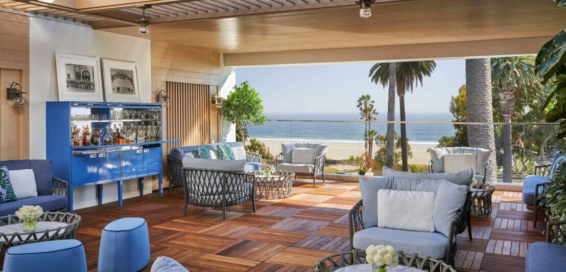 Überdachte Terrasse mit Holzboden und Polstermöbeln in Blautönen mit Blick auf Strand und Palmen