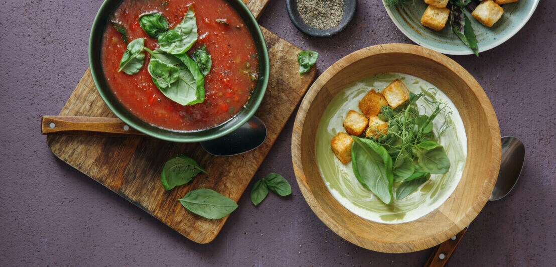 Aufsicht einer roten und einer grünen Gemüsesuppe, garniert mit Croutons und Kräutern, in Schalen auf einem Tisch