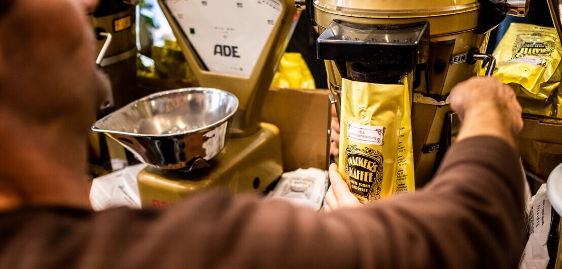 Eine Person von hinten in einem Kaffeegeschäft vor einer mechanischen Waage bei der Abfüllung von Bohnen in eine gelbe Verpackung