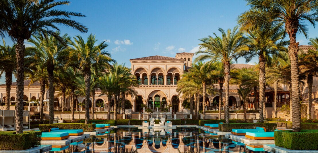 Ein luxuriöser Hotelpool, in dem sich Palmen, Sonnenliegen und das Hotelgebäude spiegeln