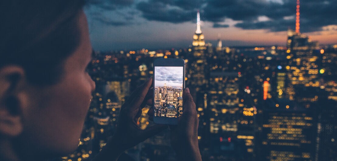 Eine Frau hält ein Smartphone in der Hand und fotografiert die nächtliche Skyline einer Stadt