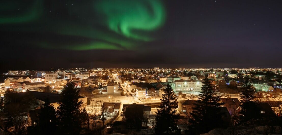 Grünes Polarlicht am Nachthimmel über einer beleuchteten Stadt