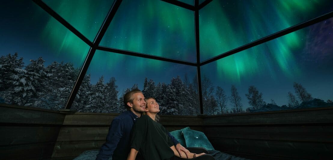 Ein Paar sitzt in einem Glas-Iglu unter einem Himmel mit Polarlichtern