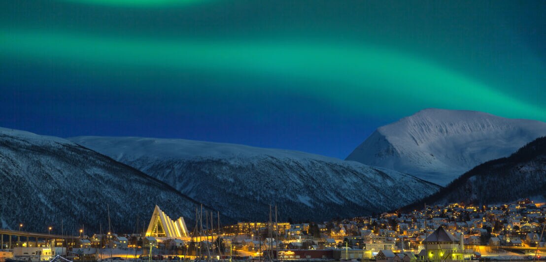 Eine beleuchtete Stadt bei Nacht vor schneebedeckten Bergen und grün schimmernden Polarlichtern am Himmel