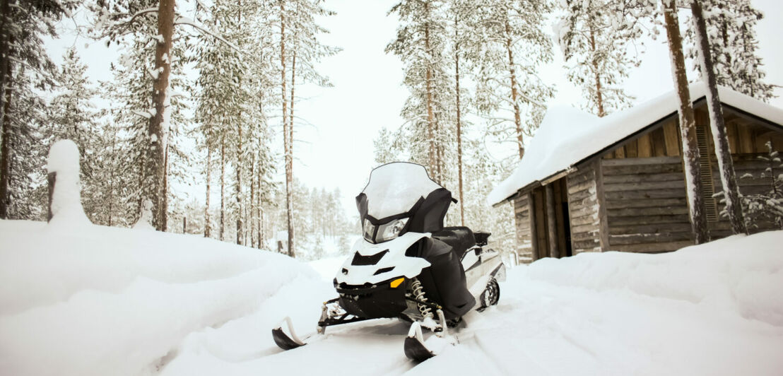 Ein Schneemobil steht auf einem schneebedeckten Weg vor einer kleinen Holzhütte