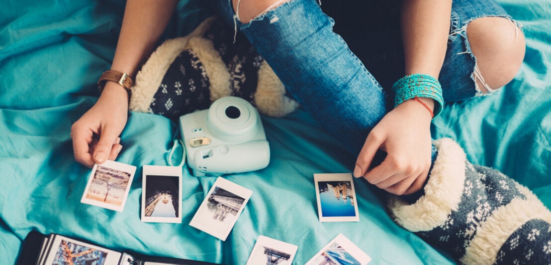 Eine junge Person, vor der eine Sofortbildkamera und ein Fotoalbum liegen, sortiert mehrere Fotos auf einem Bett