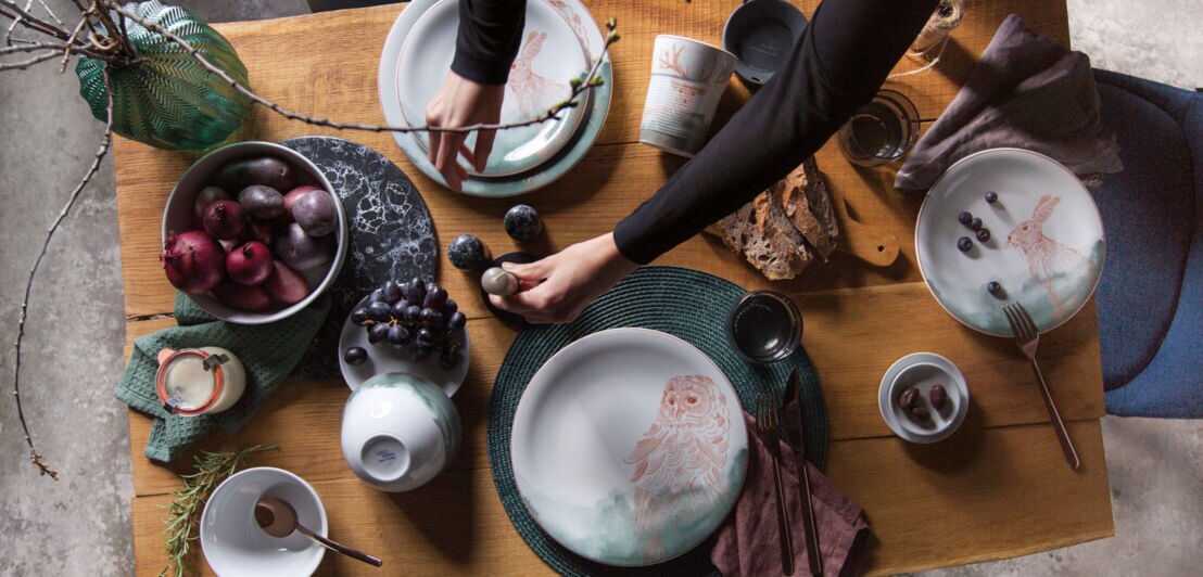 Ein gedeckter Holztisch mit kunstvoll bemaltem Porzellangeschirr, Brot, Obst und einer Vase mit Ästen