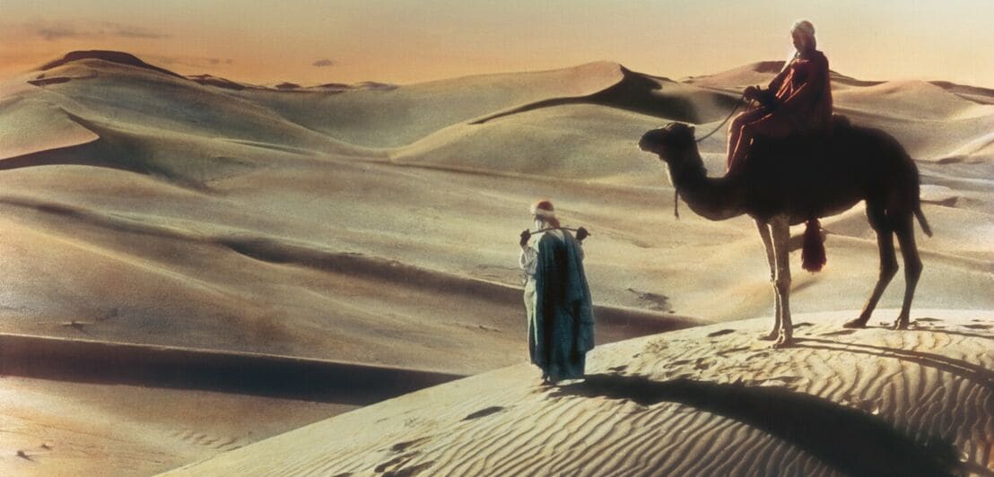 Historisches, handkoloriertes Foto von zwei Nomaden mit Kamel in der Wüste bei Sonnenuntergang