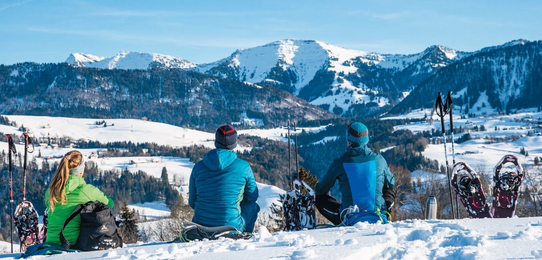 Drei Menschen in Winterkleidung sitzen im Schnee und blicken in die Ferne