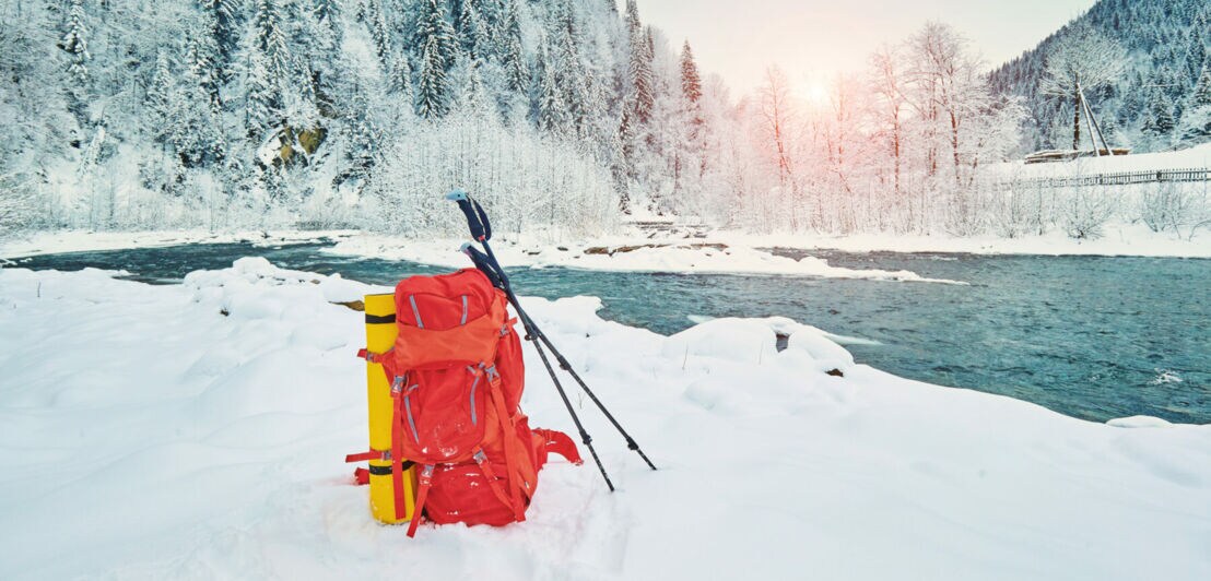 An einem Fluss steht ein Rucksack mit an ihm befestigter Matte und angelehnten Wanderstöcken im Schnee